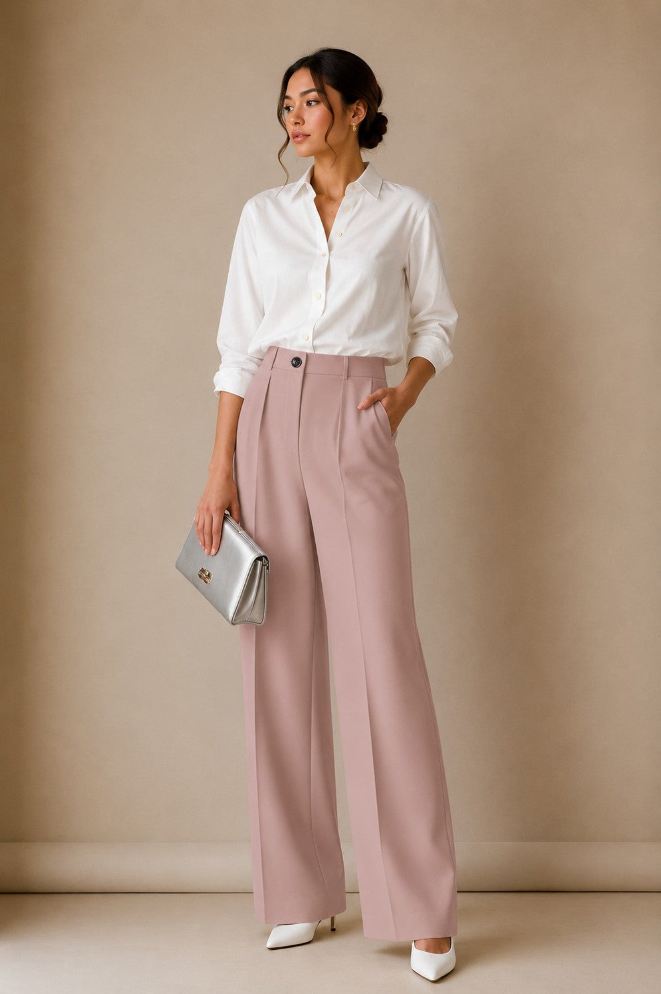 A woman stands in a studio wearing a white button-up shirt, dusty pink wide-leg trousers, and white pointed shoes, holding a metallic silver clutch against a warm beige backdrop.