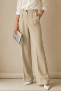 A woman stands against a warm beige studio backdrop wearing a white button-up blouse, high-waisted wide-leg beige trousers, white pointed shoes, and holding a metallic silver clutch.