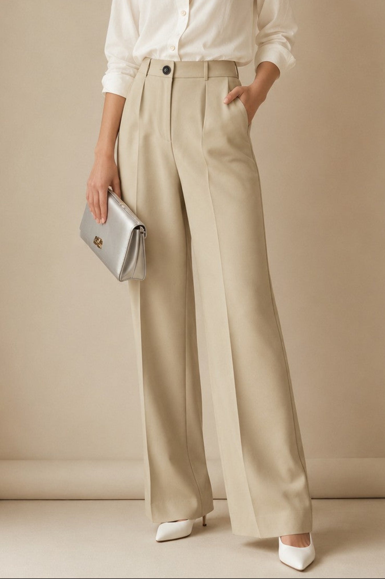A woman stands against a warm beige studio backdrop wearing a white button-up blouse, high-waisted wide-leg beige trousers, white pointed shoes, and holding a metallic silver clutch.