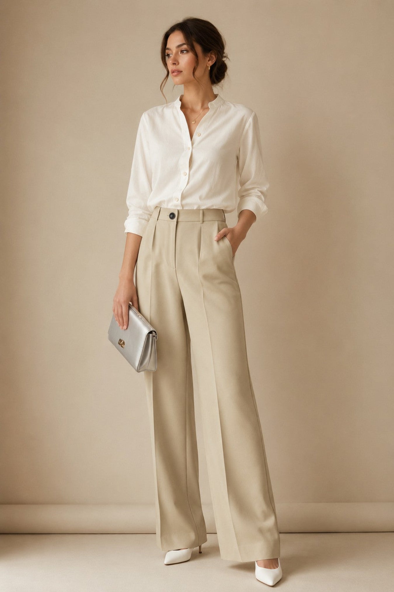 A woman stands against a warm beige studio backdrop wearing a white button-up blouse, high-waisted wide-leg beige trousers, white pointed shoes, and holding a silver clutch.