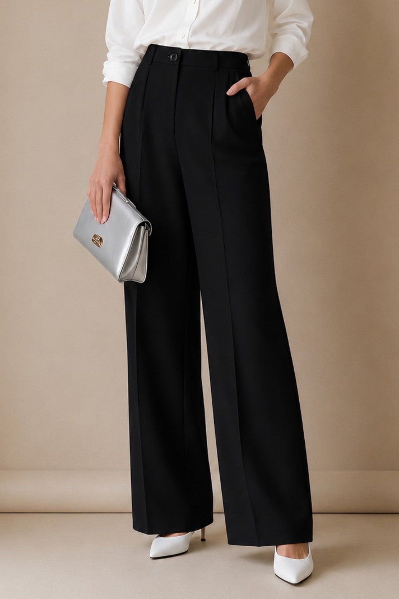 A woman stands against a beige studio wall wearing a white button-up blouse, black wide-leg trousers, white pointed heels, and holding a silver clutch.