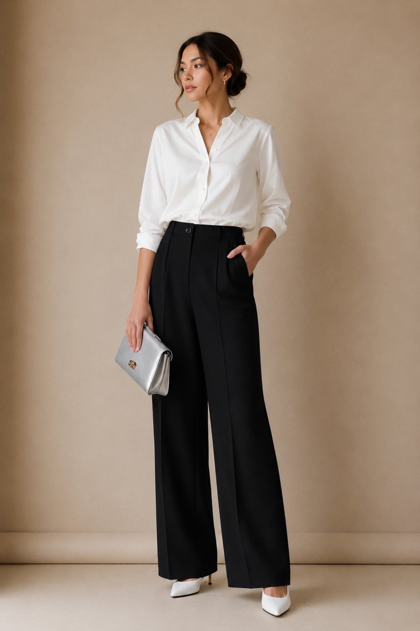 A woman stands against a beige studio wall wearing a white button-up blouse, black wide-leg trousers, white pointed heels, and holding a silver clutch.