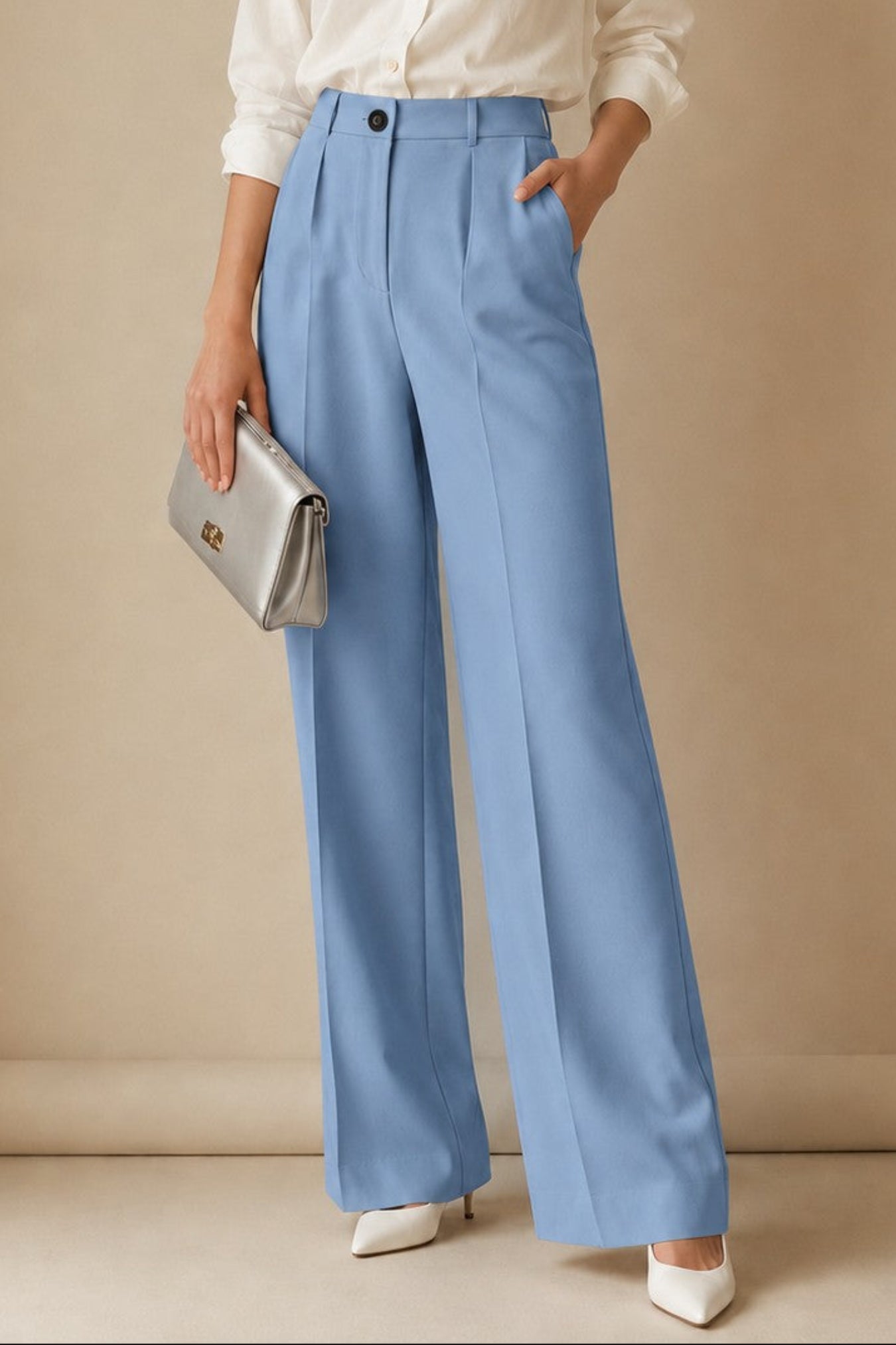 A woman stands against a warm beige studio backdrop wearing a white button-up blouse, light blue wide-leg trousers, and white pointed shoes, while holding a metallic silver clutch.