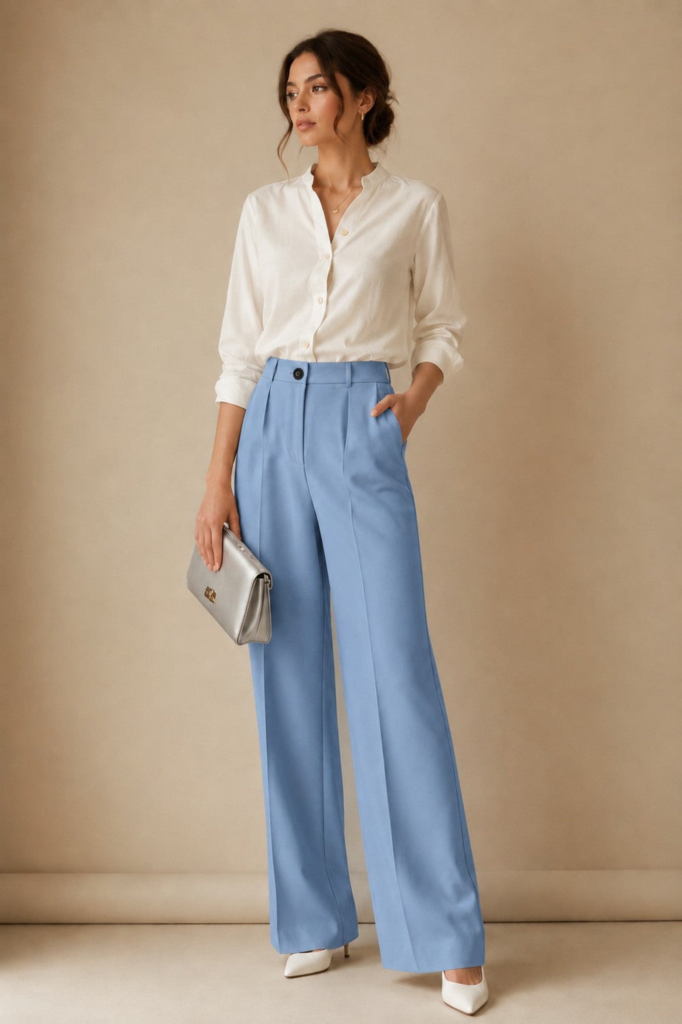 A woman stands against a warm beige studio backdrop wearing a white button-up blouse, light blue wide-leg trousers, white pointed shoes, and holding a silver clutch.