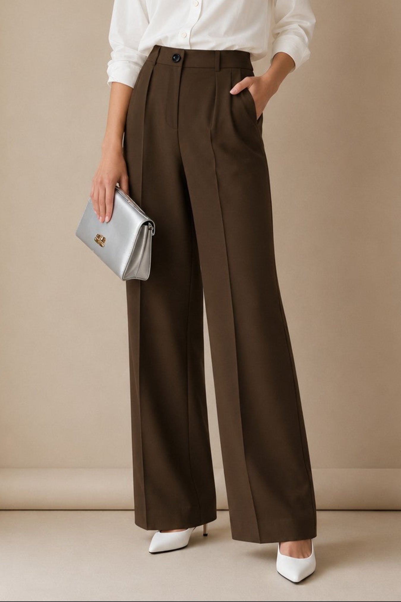 A woman stands in a studio wearing a white button-up blouse, dark brown wide-leg trousers, white pointed shoes, and holding a silver clutch.