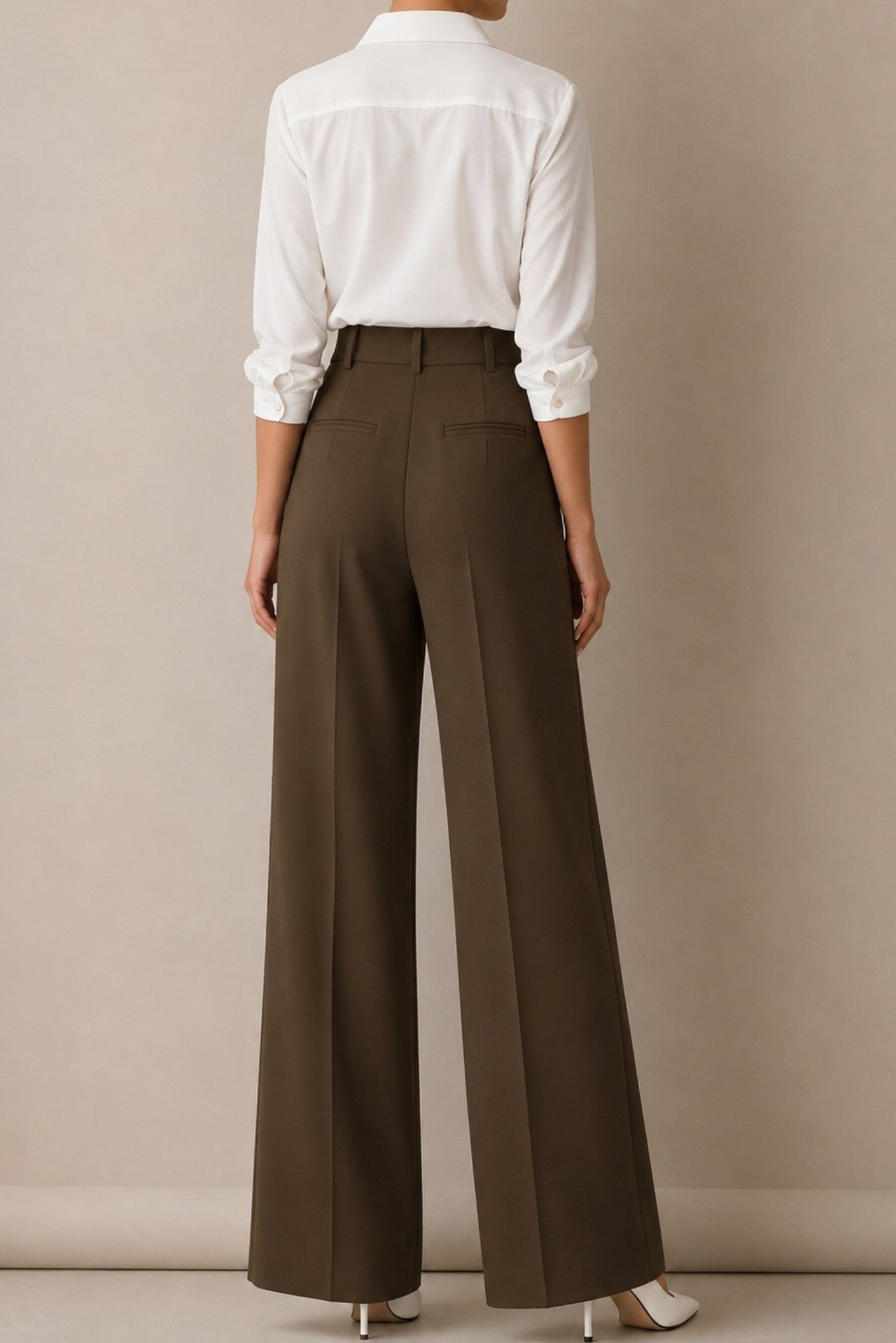 A model shown from the back wearing high-waisted, wide-leg brown trousers and a white long-sleeve button-up shirt, standing against a neutral beige studio backdrop.
