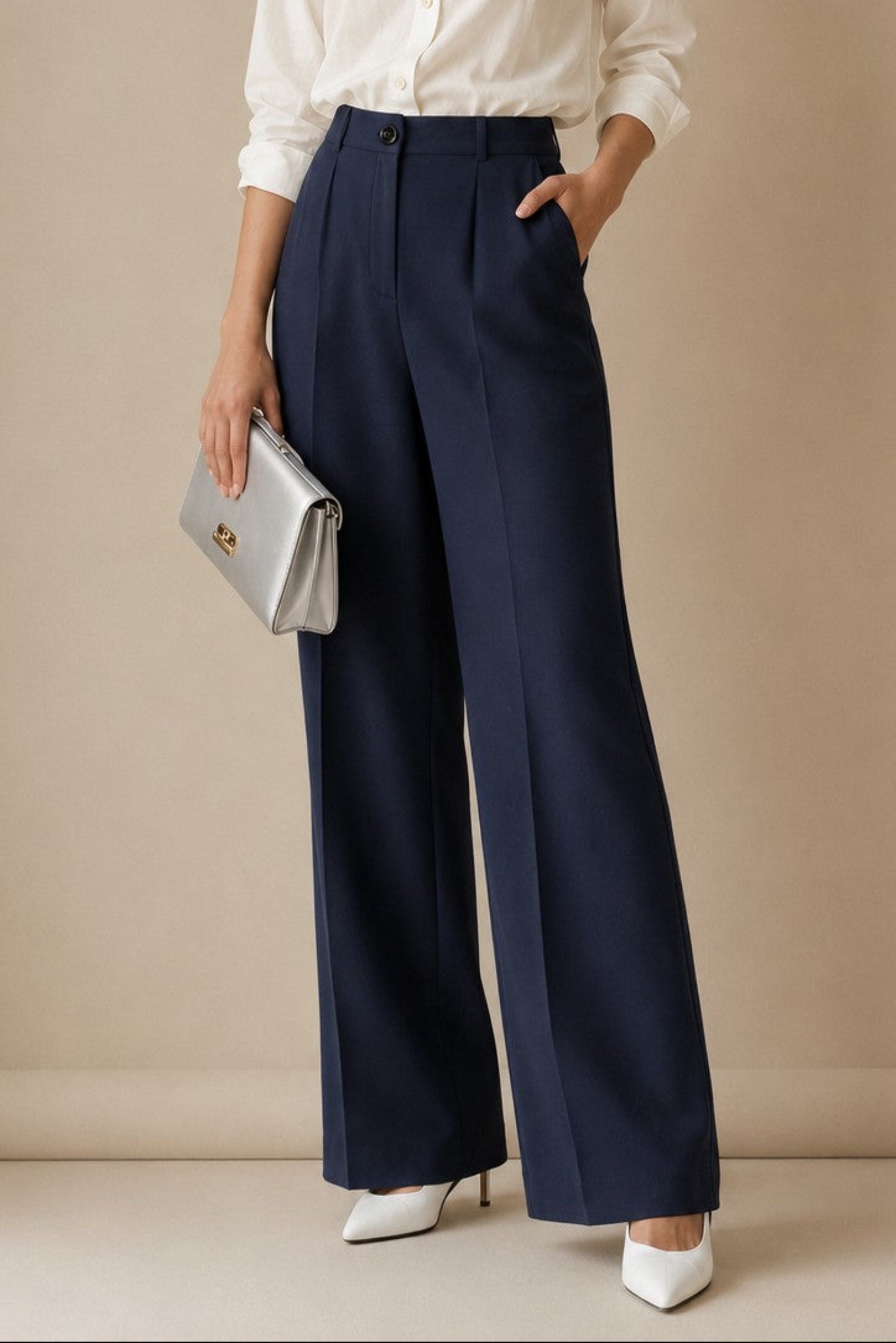 A woman stands in a studio wearing a white button-up shirt, navy wide-leg trousers, and white pointed heels, holding a small silver clutch against a warm beige backdrop.
