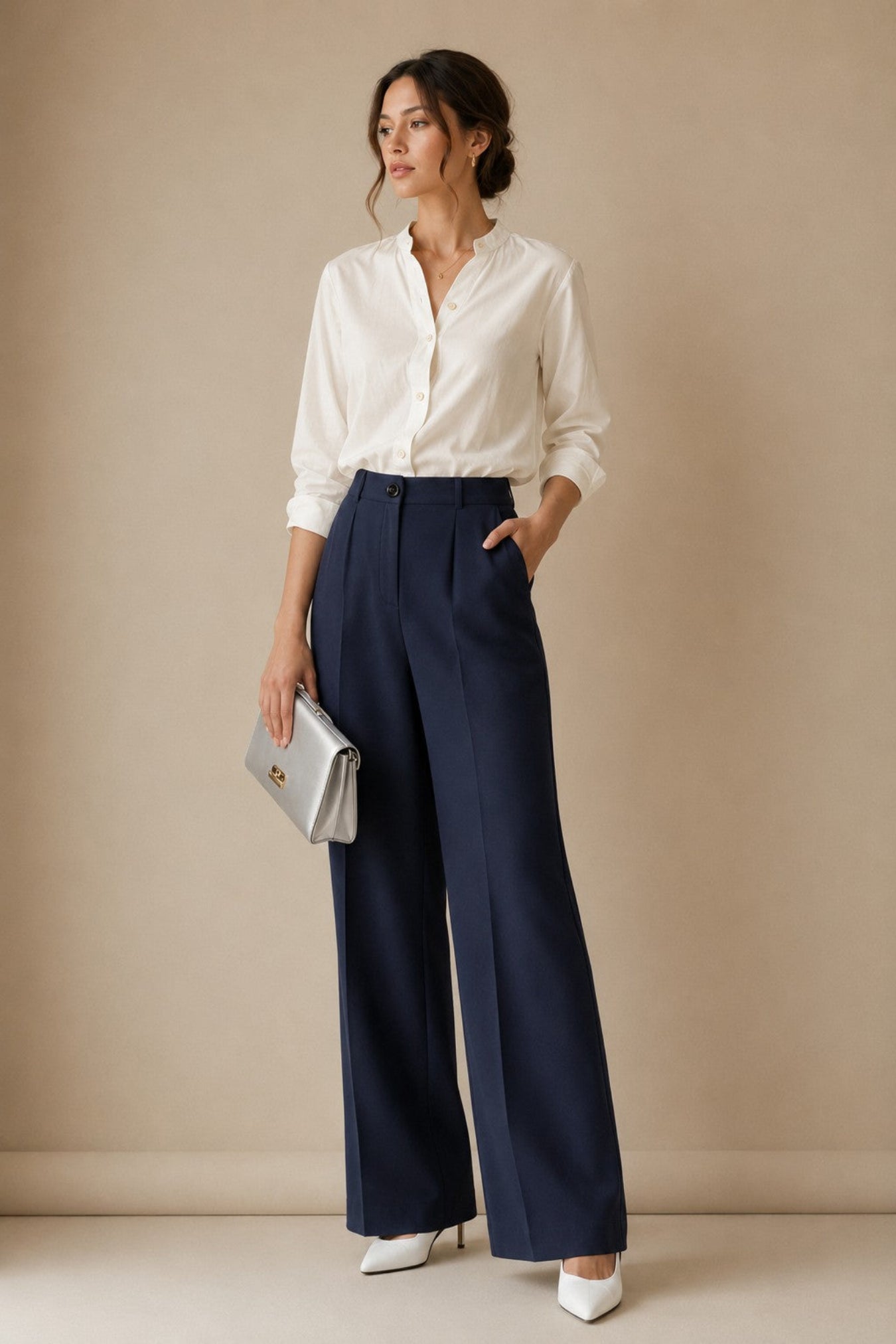 A woman stands in a studio wearing a white button-up shirt, navy wide-leg trousers, and white pointed heels, holding a small silver clutch against a warm beige backdrop.