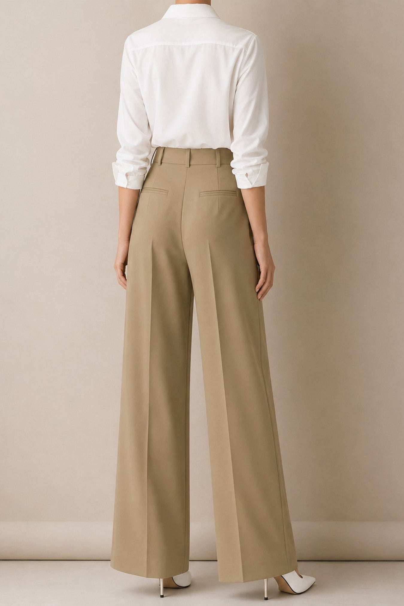 A model shown from the back wearing high-waisted, wide-leg taupe trousers and a white button-down shirt, standing against a neutral beige studio background.