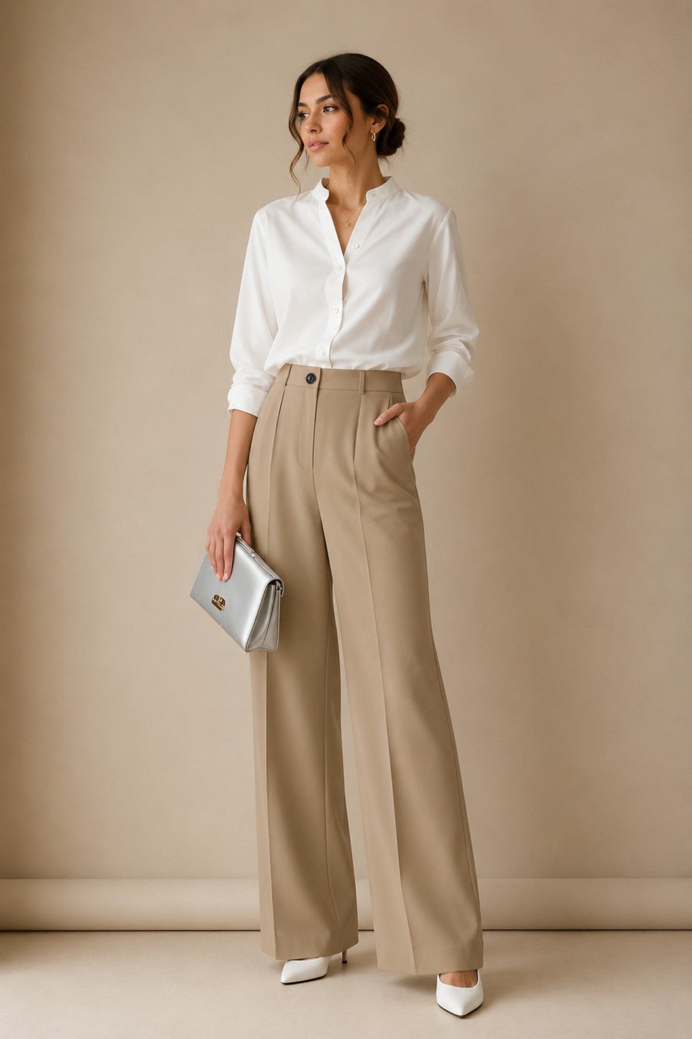 A woman stands in a studio wearing a white button-up blouse, high-waisted beige wide-leg trousers, white pointed shoes, and holding a metallic silver clutch.