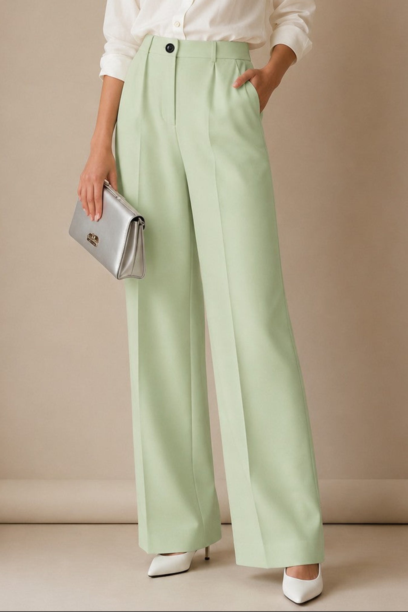 A woman stands in a studio wearing a white button-up shirt, light sage green wide-leg trousers, and white pointed shoes, holding a silver clutch against a warm beige backdrop.