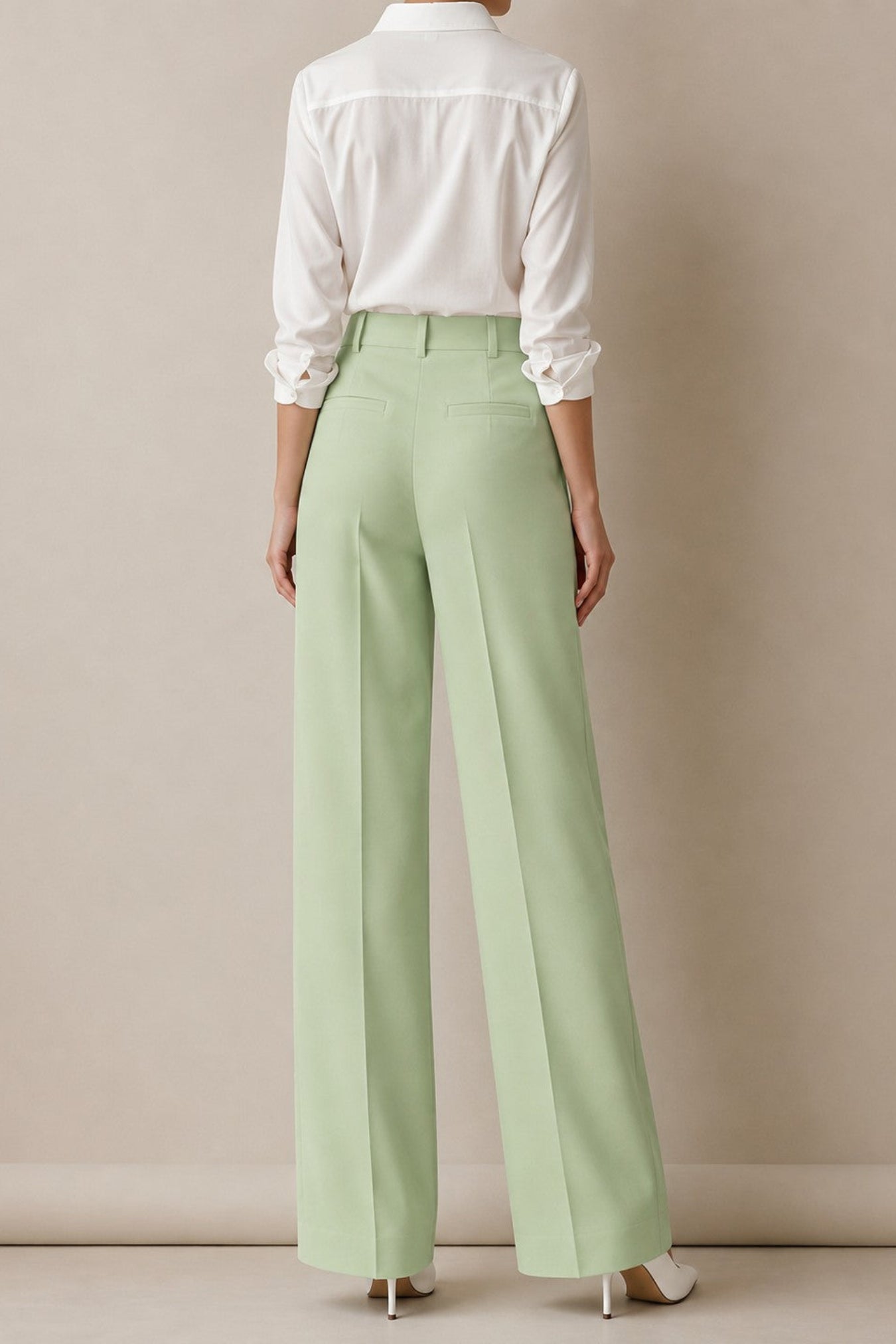 A model shown from the back wearing light sage green wide-leg trousers, a white long-sleeve button-down shirt, and white heels, set against a neutral beige studio background.