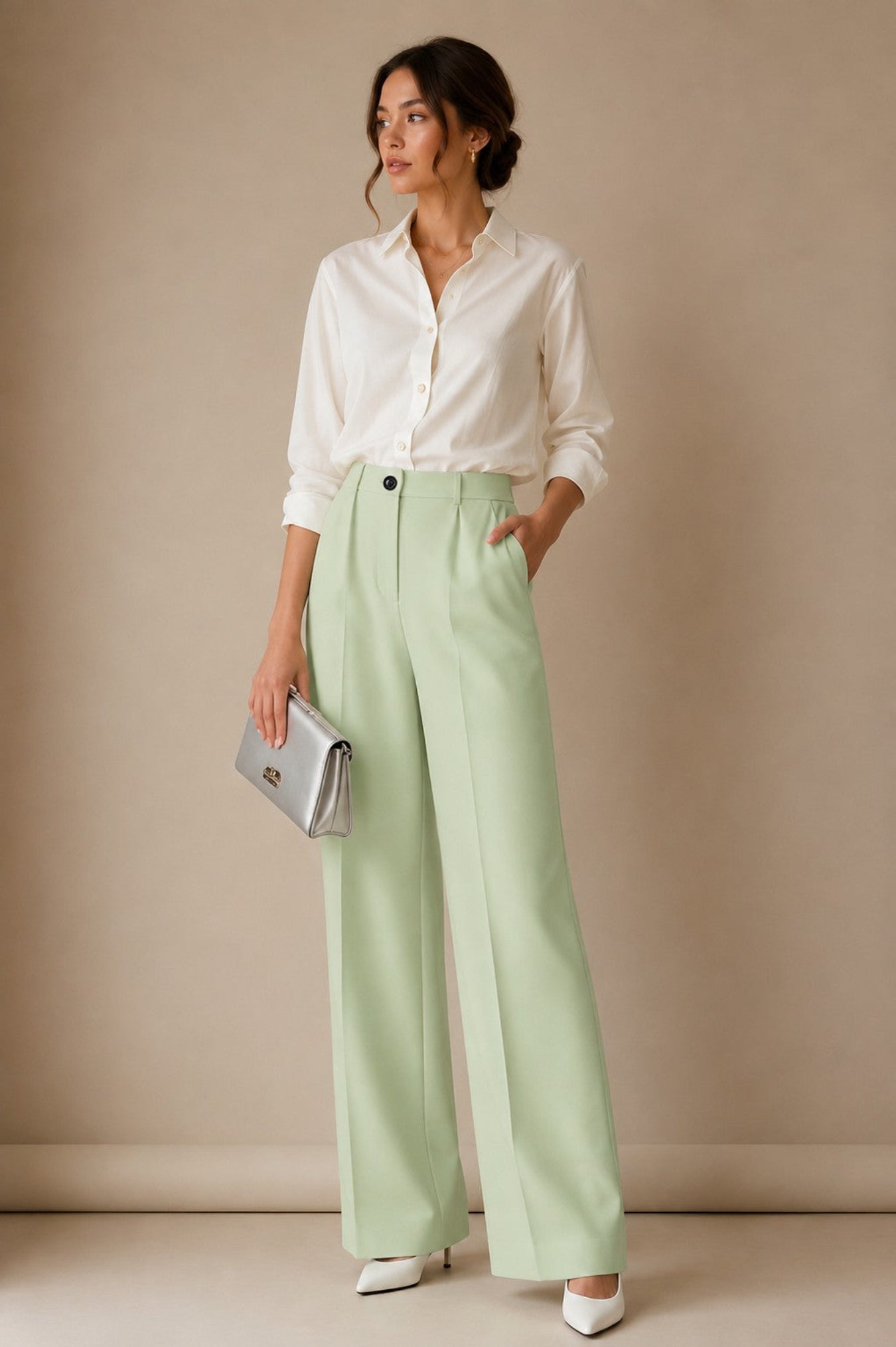 A woman stands in a studio wearing a white button-up shirt, light sage green wide-leg trousers, and white pointed shoes, holding a silver clutch against a warm beige backdrop.