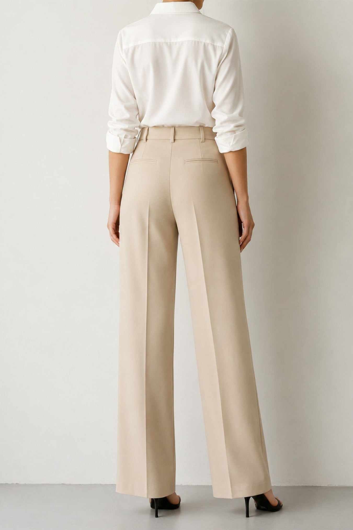 A model shown from the back wearing high-waisted, wide-leg beige trousers paired with a white button-down shirt and black heels, set against a neutral light gray studio background.