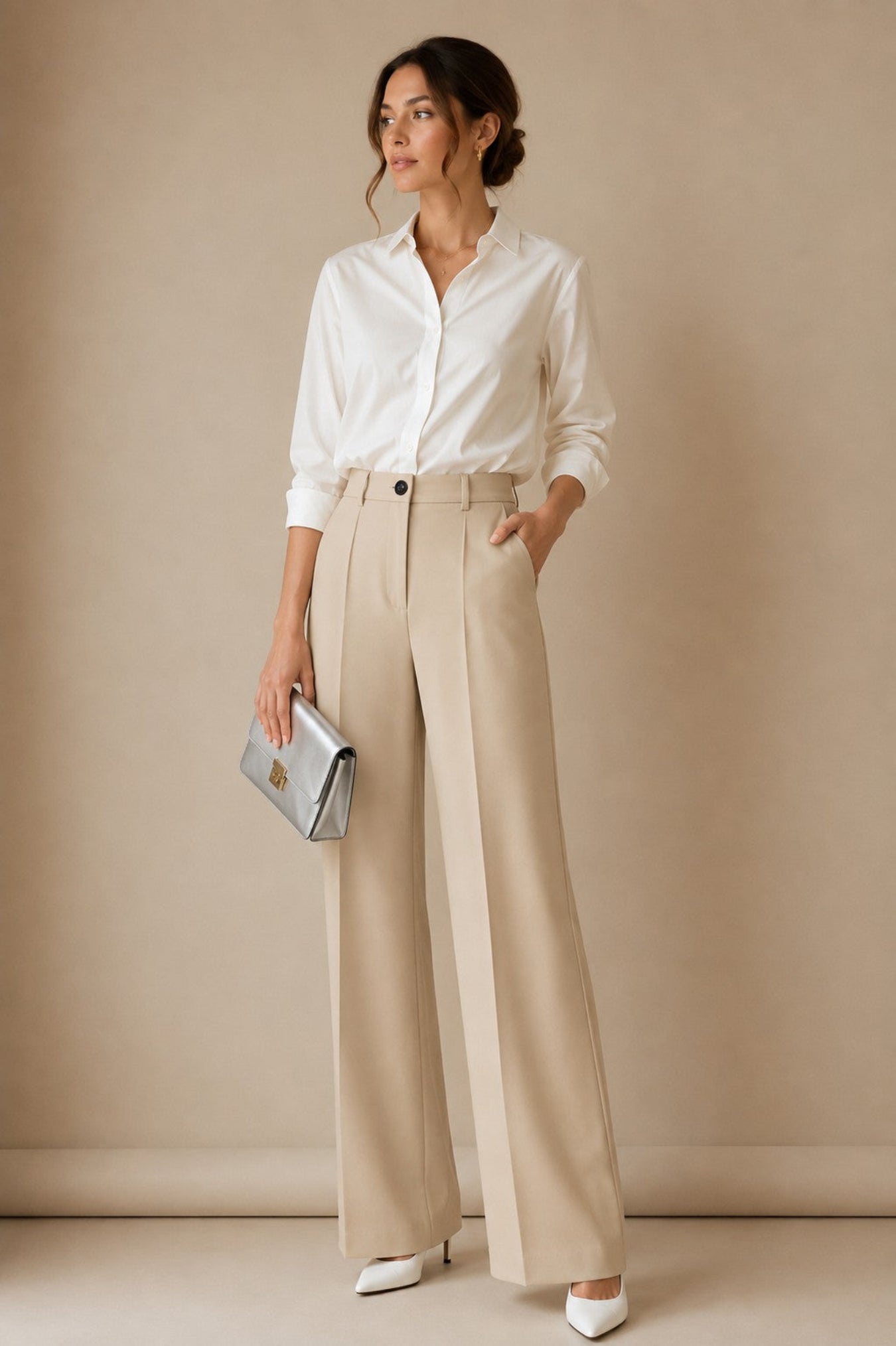 A woman stands against a warm beige studio backdrop wearing a white button-up shirt, high-waisted beige wide-leg trousers, white pointed heels, and holding a metallic silver clutch.