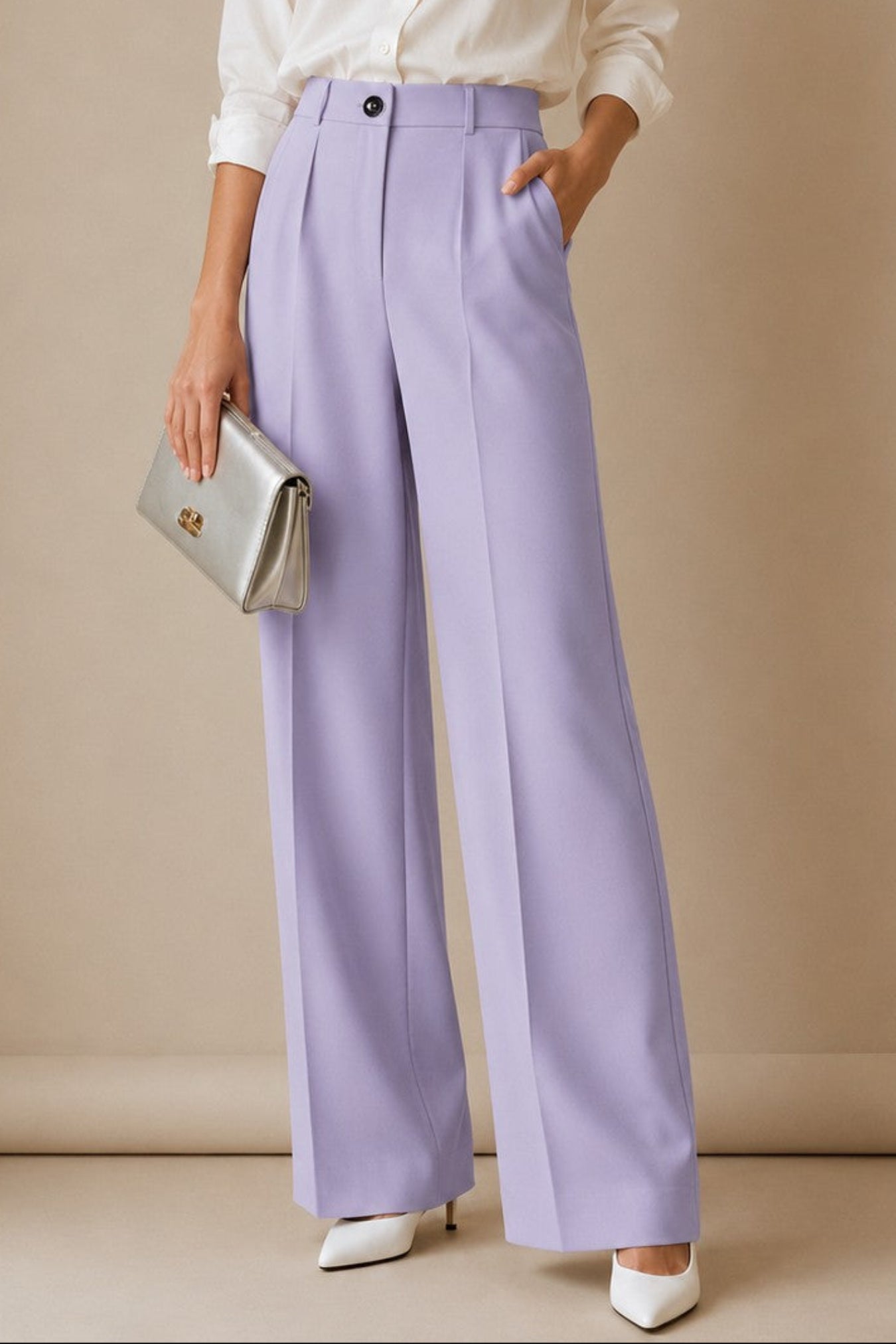A woman stands against a beige studio backdrop wearing a white button-up shirt, lavender wide-leg trousers, white pointed shoes, and holding a silver clutch.