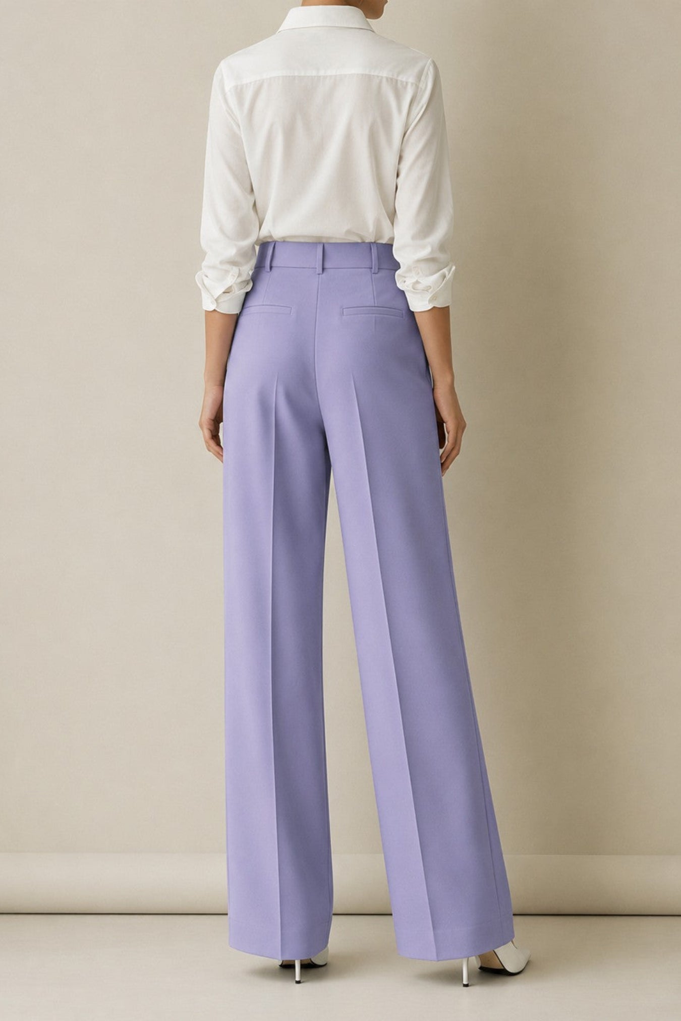 A model shown from the back wearing lavender wide-leg trousers, a white long-sleeve button-up shirt, and white pointed-toe heels against a neutral beige studio background.