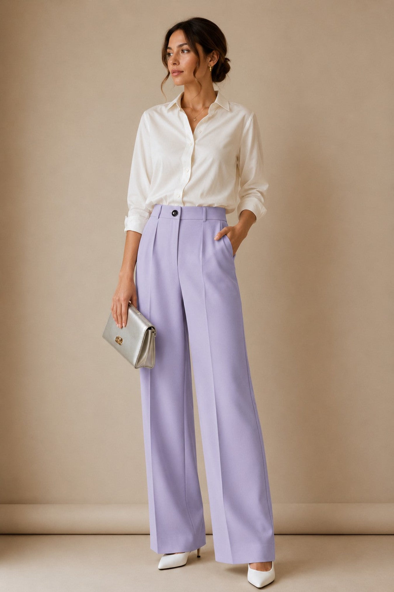 A woman stands against a warm beige studio backdrop wearing a white button-up shirt, lavender wide-leg trousers, white pointed heels, and holding a metallic silver clutch.