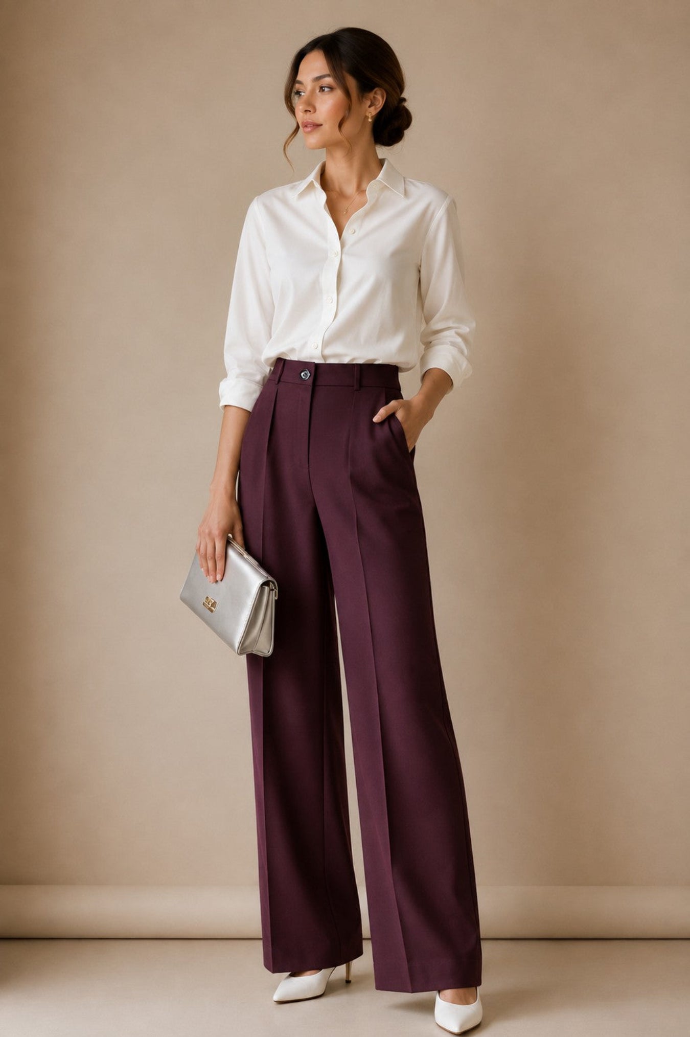 A woman stands in a studio wearing a white button-up shirt, deep plum wide-leg trousers, white heels, and holding a silver clutch. The background is a solid warm beige studio wall.