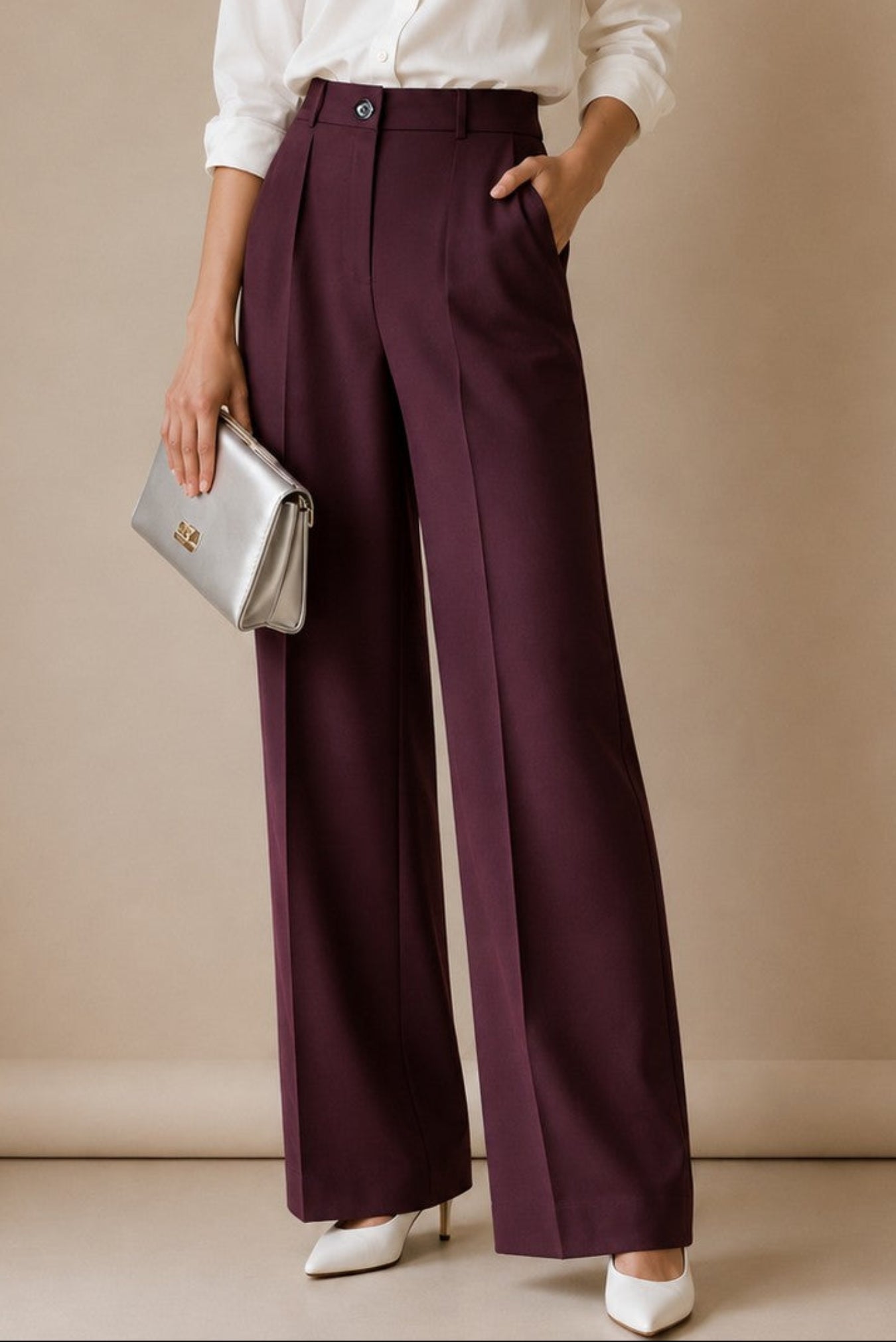 A woman stands in a studio wearing a white button-up shirt, deep plum wide-leg trousers, white heels, and holding a silver clutch. The background is a solid warm beige studio wall.