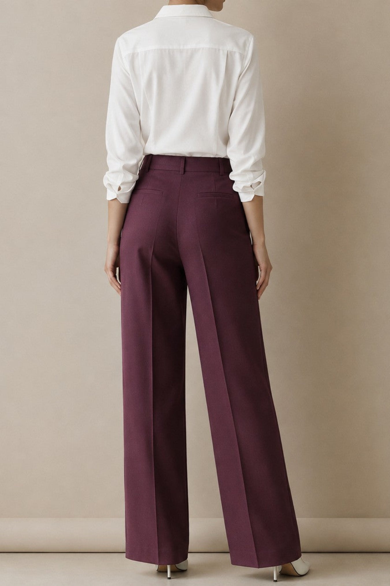 A model shown from the back wearing high-waisted, wide-leg trousers in a deep plum purple, paired with a white long-sleeve button-down shirt. The setting is a clean studio with a warm beige background.