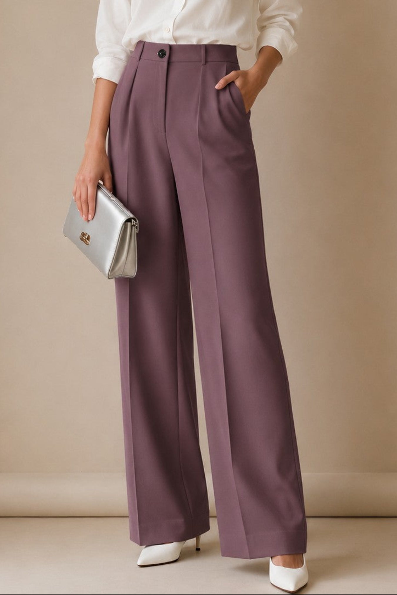 A woman stands in a studio wearing a white button-up shirt, mauve wide-leg trousers, white pointed shoes, and holding a silver clutch.