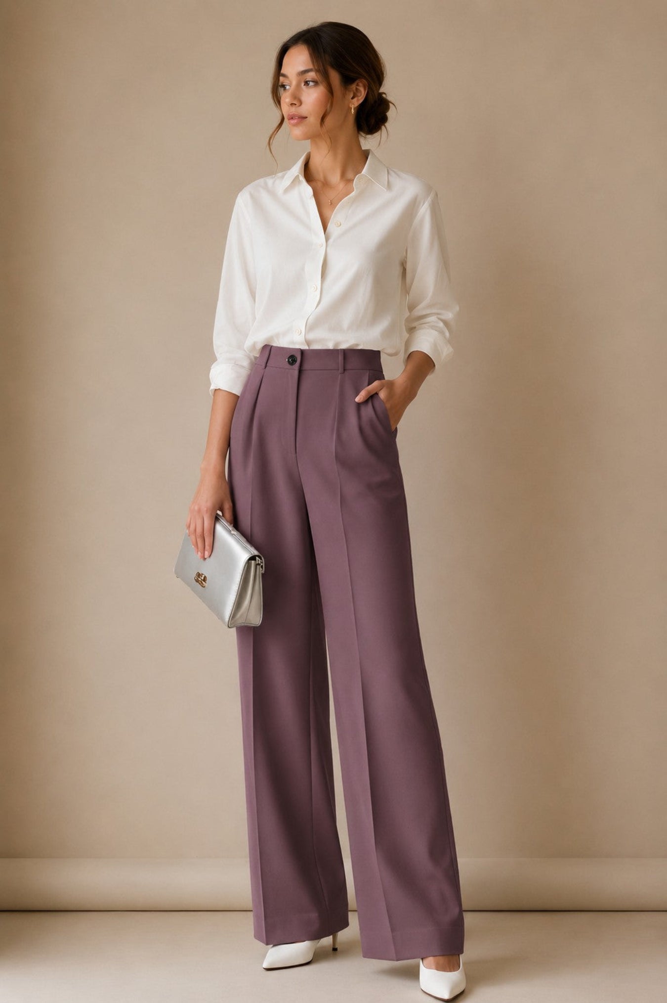 A woman stands in a studio wearing a white button-up shirt, mauve wide-leg trousers, white pointed shoes, and holding a silver clutch.