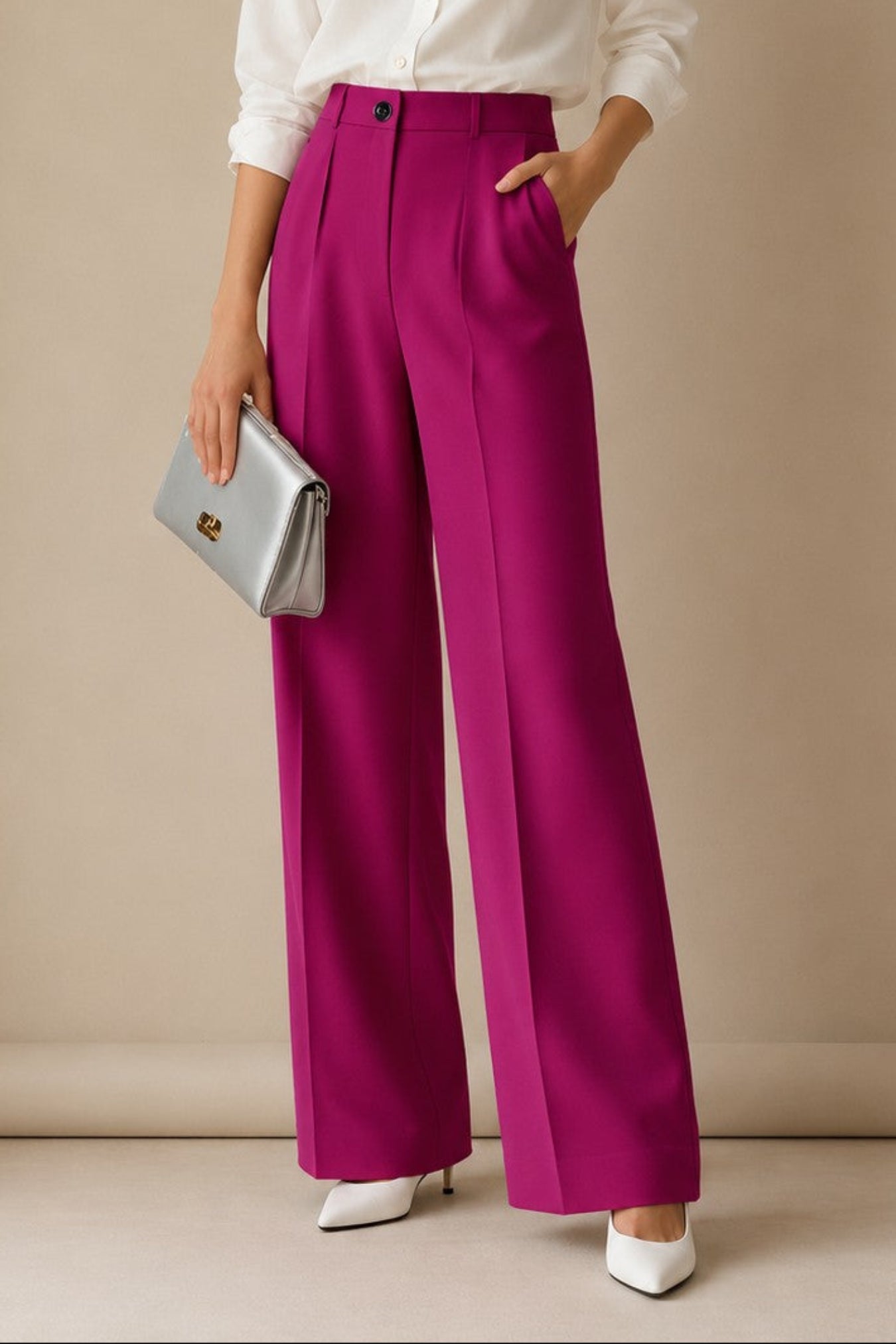 A woman stands against a beige studio wall wearing a white button-up blouse, bright magenta wide-leg trousers, white pointed shoes, and holding a silver clutch.