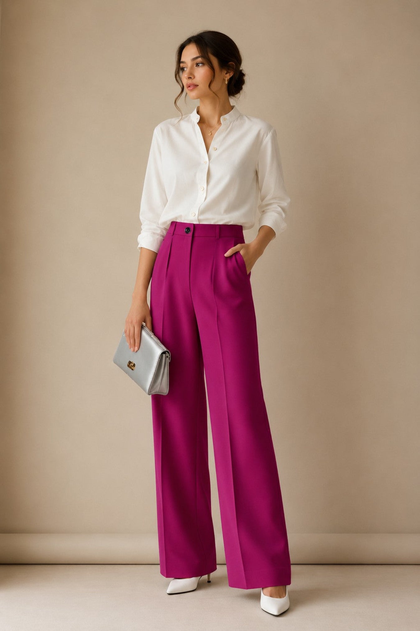 A woman stands against a beige studio wall wearing a white button-up blouse, bright magenta wide-leg trousers, white pointed shoes, and holding a silver clutch.