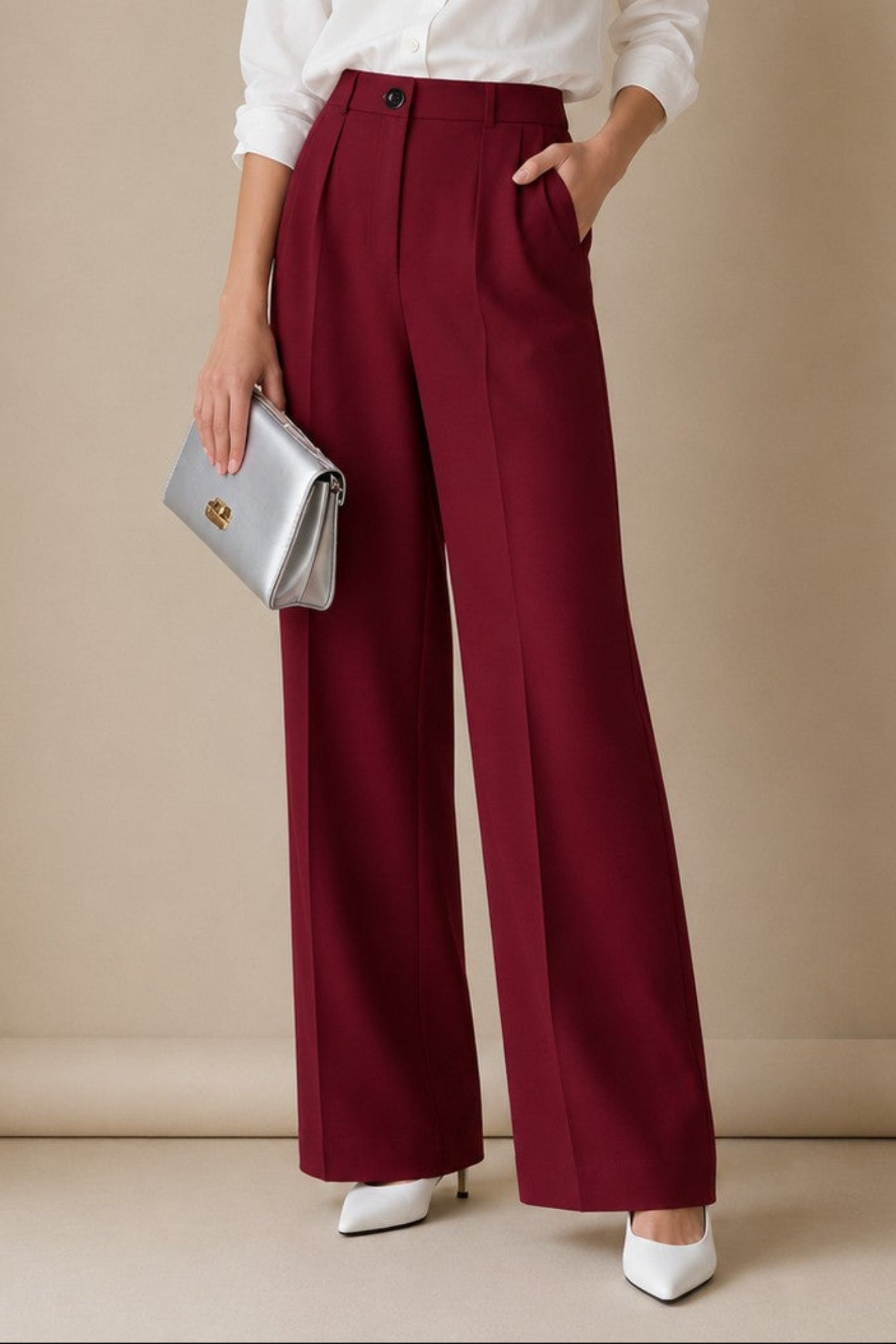 A woman stands against a beige studio backdrop wearing a white button-up blouse, high-waisted burgundy wide-leg trousers, and white pointed heels, while holding a small metallic clutch.