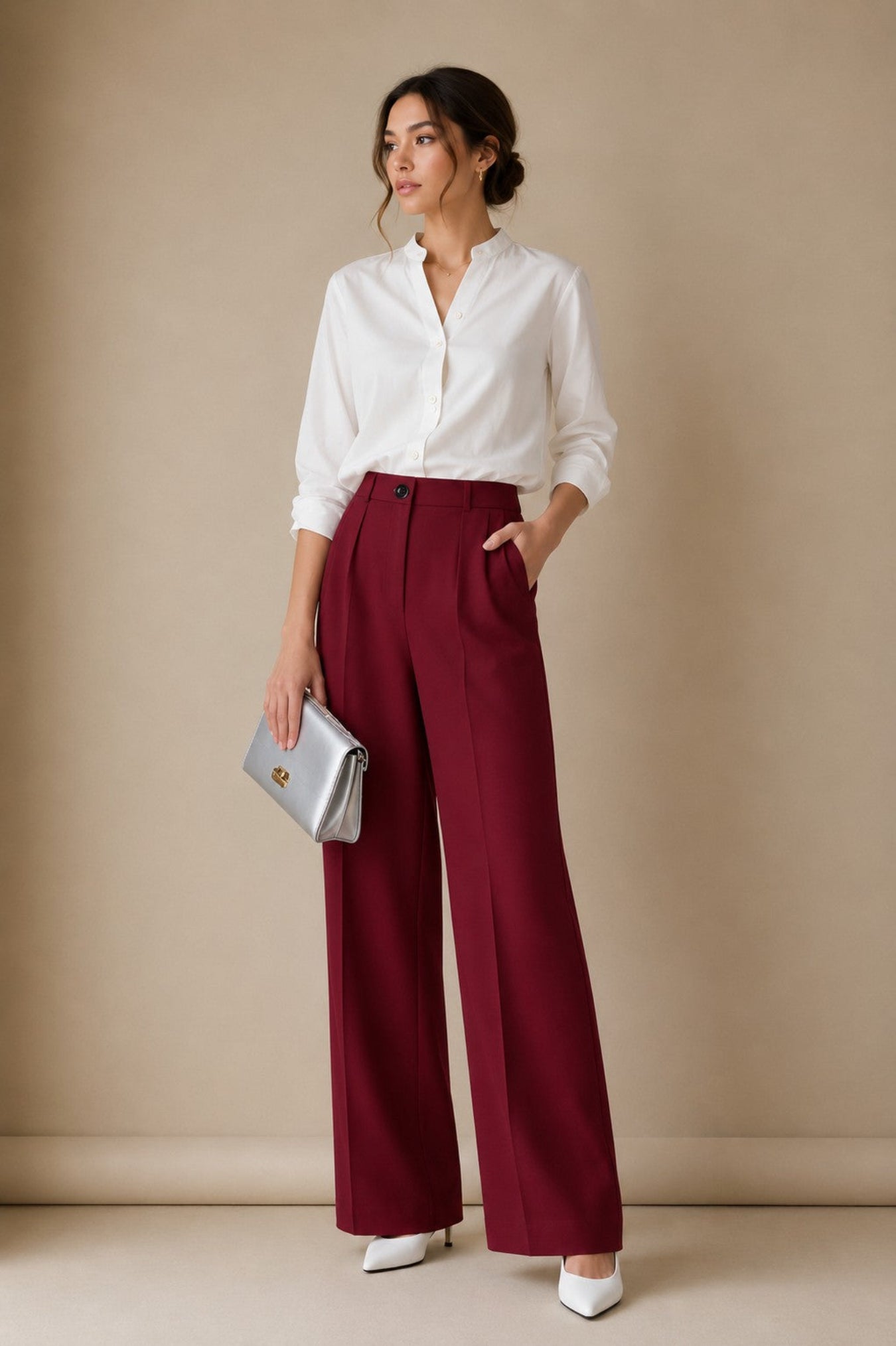 A woman stands in a studio wearing a white button-up blouse, high-waisted burgundy wide-leg trousers, white pointed heels, and holding a silver clutch bag against a warm beige backdrop.