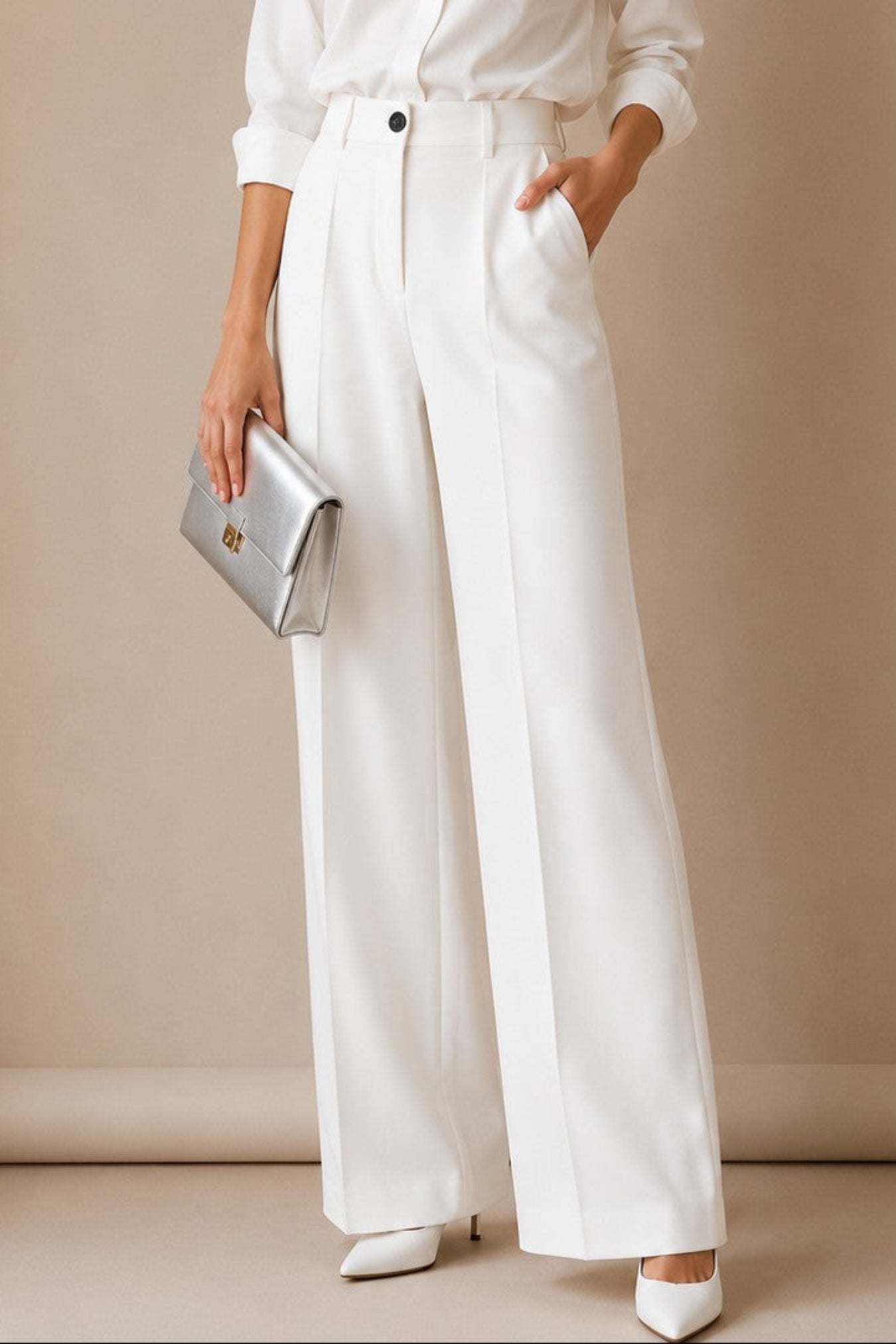 A woman models a white long-sleeve button-up shirt and matching high-waisted white wide-leg trousers, holding a metallic silver clutch against a neutral beige studio backdrop.