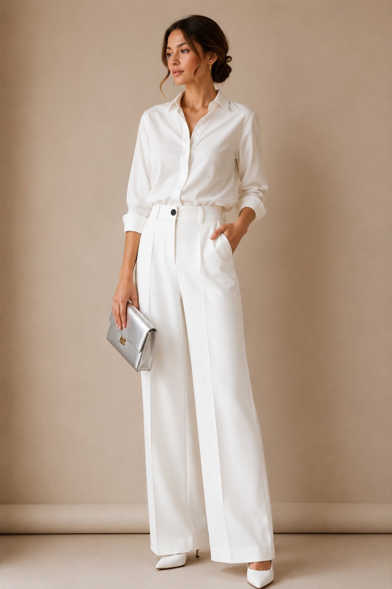 A woman modeling a white long-sleeve button-up shirt and matching white wide-leg trousers with a tie waist, holding a metallic silver clutch against a warm beige studio backdrop.
