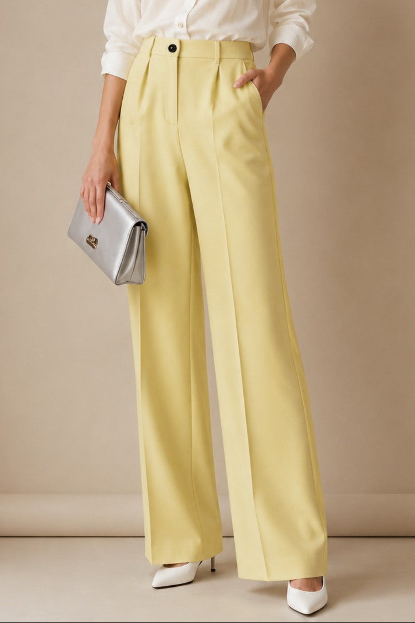 A woman stands against a warm beige studio backdrop wearing a white button-up shirt, pale yellow wide-leg trousers, white pointed shoes, and holding a silver clutch.