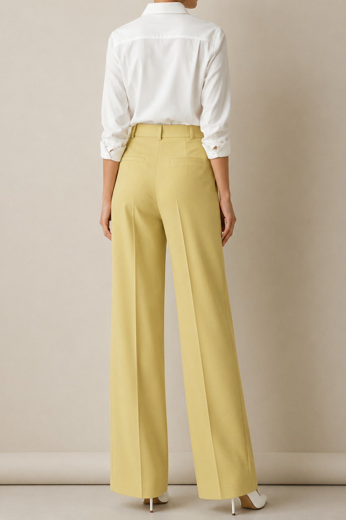 A model shown from the back wearing high-waisted, wide-leg trousers in a pale yellow/ochre color, paired with a white long-sleeve button-down shirt and white heels, against a neutral beige studio background.