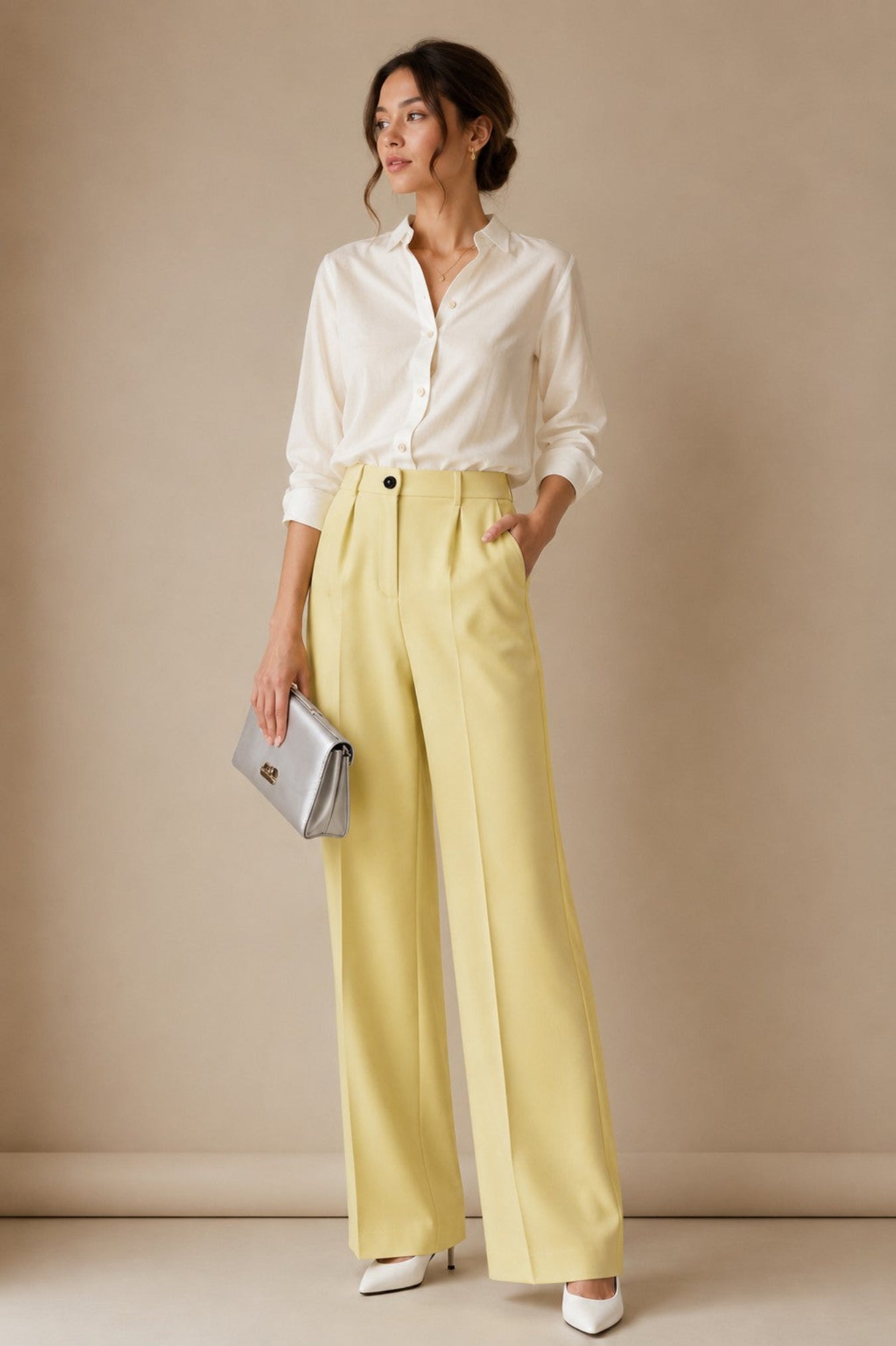 A woman stands against a warm beige studio backdrop wearing a white button-up shirt, pale yellow wide-leg trousers, white pointed shoes, and holding a silver clutch.