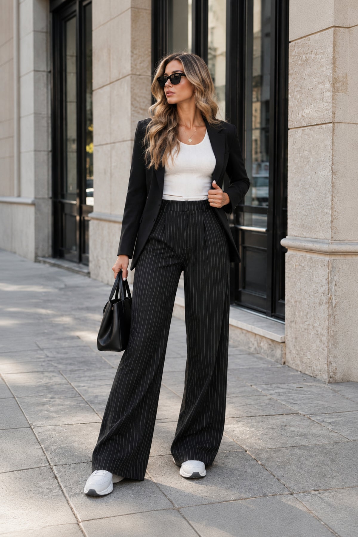 A woman walking on a city sidewalk wearing a black pinstripe wide-leg blazer and matching pinstripe trousers over a white tank top, carrying a black handbag.