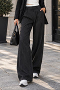 A woman stands on a stone sidewalk wearing a black pinstripe wide-leg blazer and matching pinstripe trousers, a white crop top, black sunglasses, and white sneakers, while holding a black handbag.