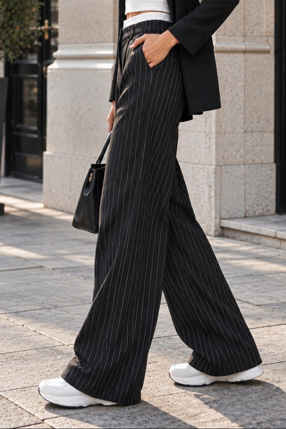 A woman walking outdoors on a city sidewalk wearing a black pinstripe wide-leg blazer and matching wide-leg trousers over a white crop top, with white sneakers and a black handbag.