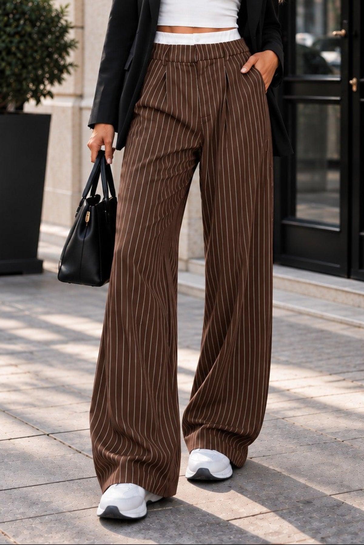 A woman stands on a city sidewalk wearing a black blazer, white crop top, and brown pinstriped wide-leg pants, with white sneakers and a black handbag.