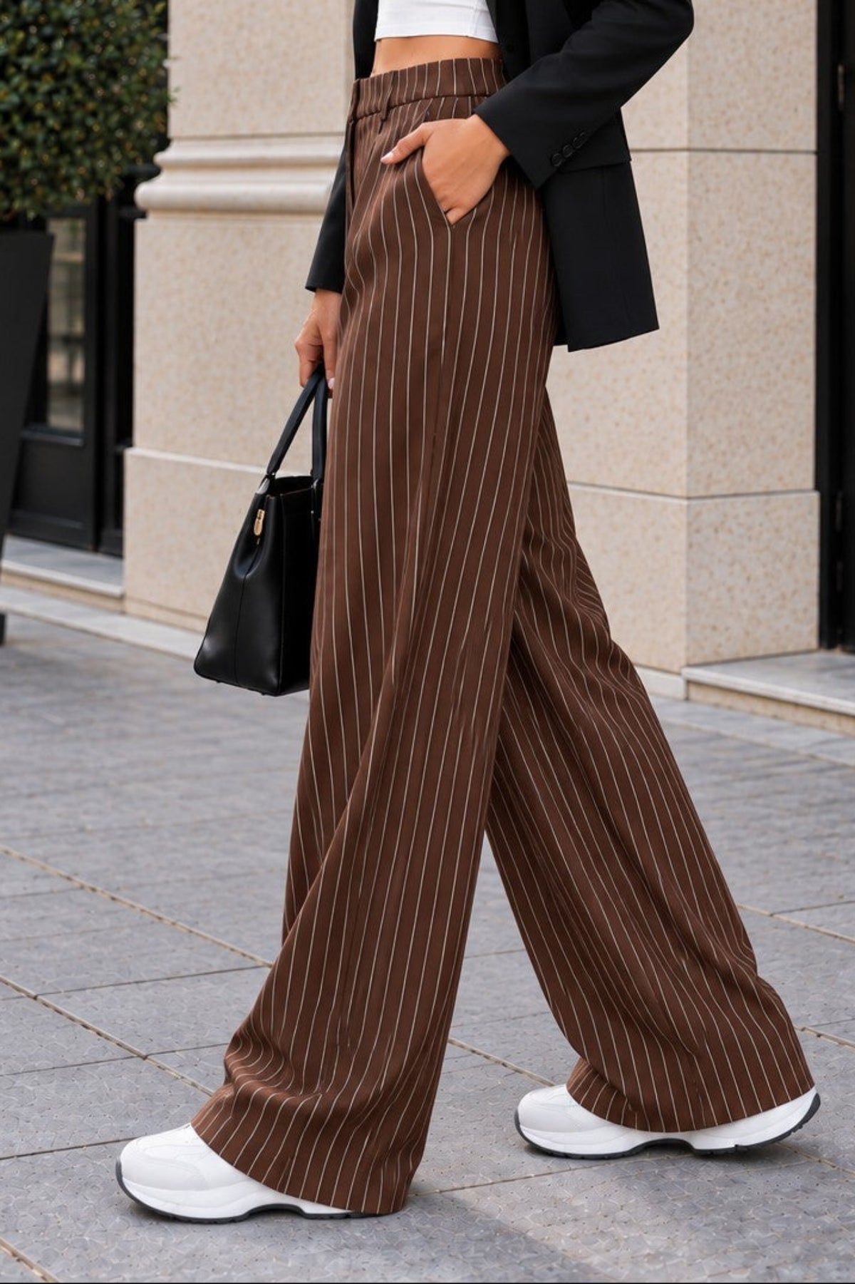 A woman walking outdoors in front of a beige stone building, wearing brown wide-leg pinstripe trousers, a white crop top, a black blazer, white sneakers, and holding a black handbag.