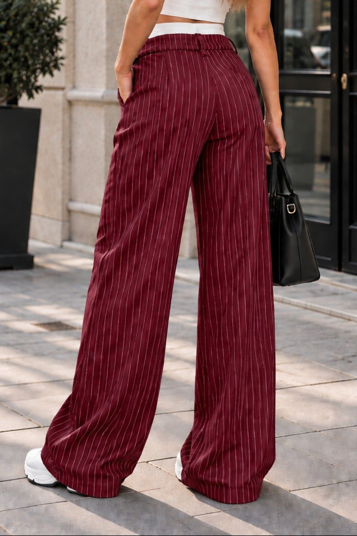 A woman stands on a city sidewalk wearing high-waisted, wide-leg burgundy trousers with thin vertical gold pinstripes, paired with a white crop top and sunglasses.