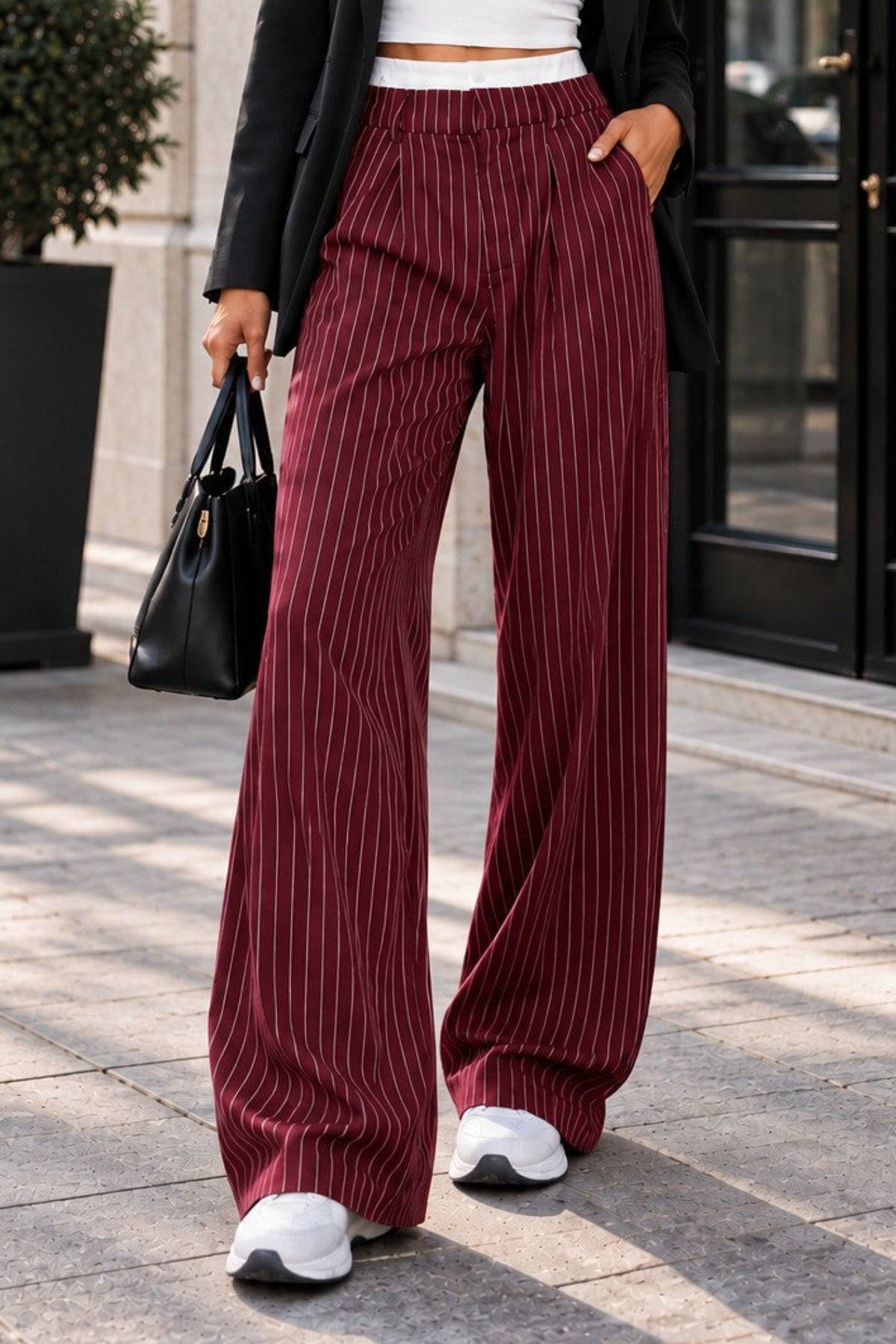 A woman stands on a city sidewalk wearing a black blazer, white crop top, and high-waisted burgundy pinstriped wide-leg pants, holding a black handbag.