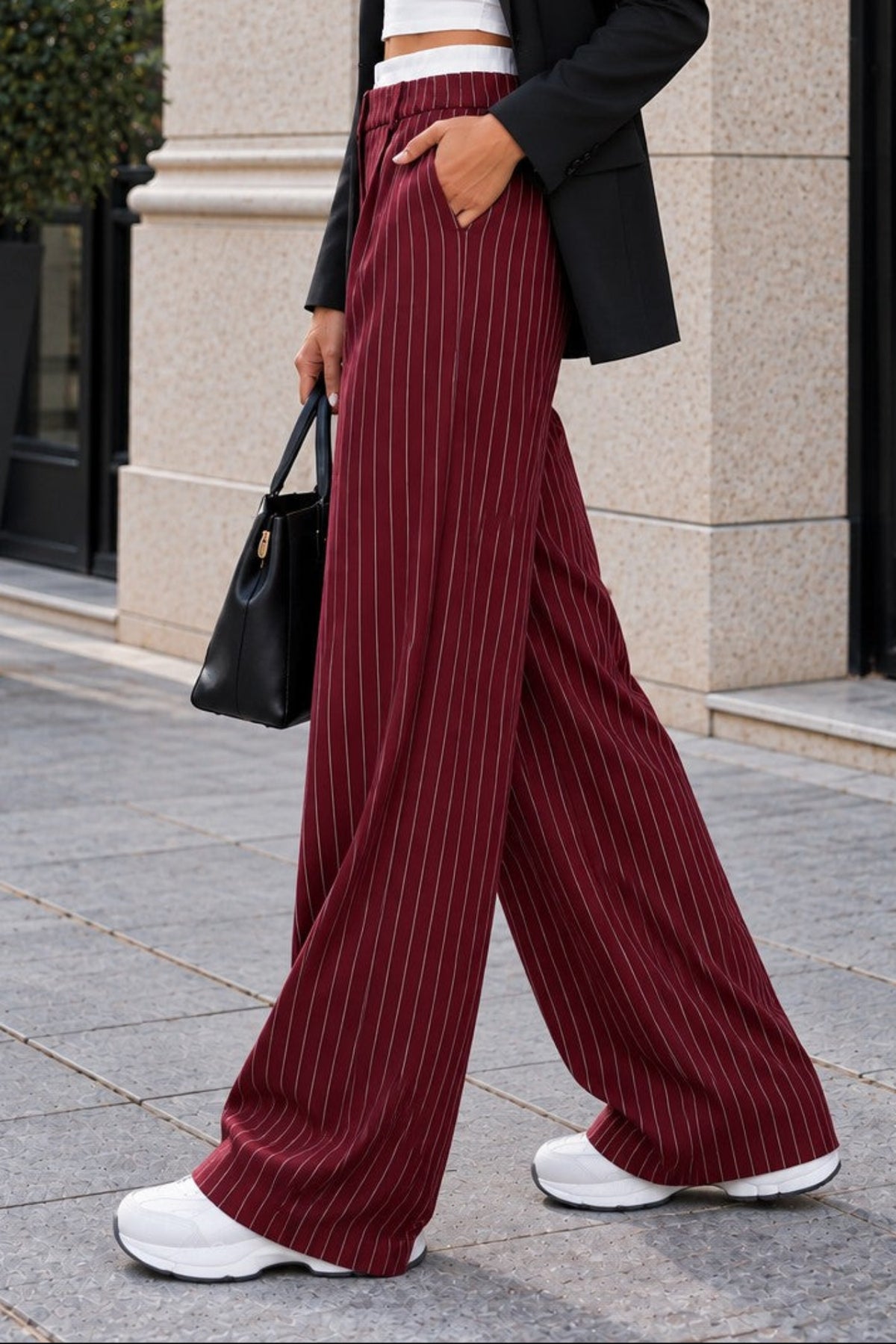 A woman walking outdoors wearing a black blazer, white crop top, and burgundy pinstriped wide-leg trousers, with white sneakers and a black handbag.