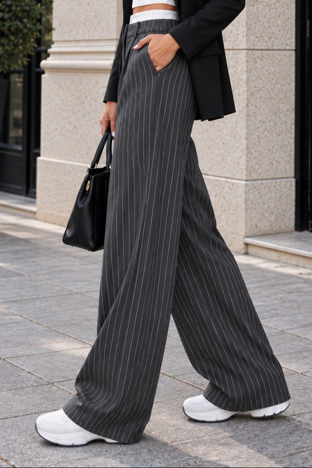 A woman walking outdoors in front of a modern building, wearing a black blazer, white crop top, black-and-white pinstripe wide-leg trousers, white sneakers, and holding a black handbag.