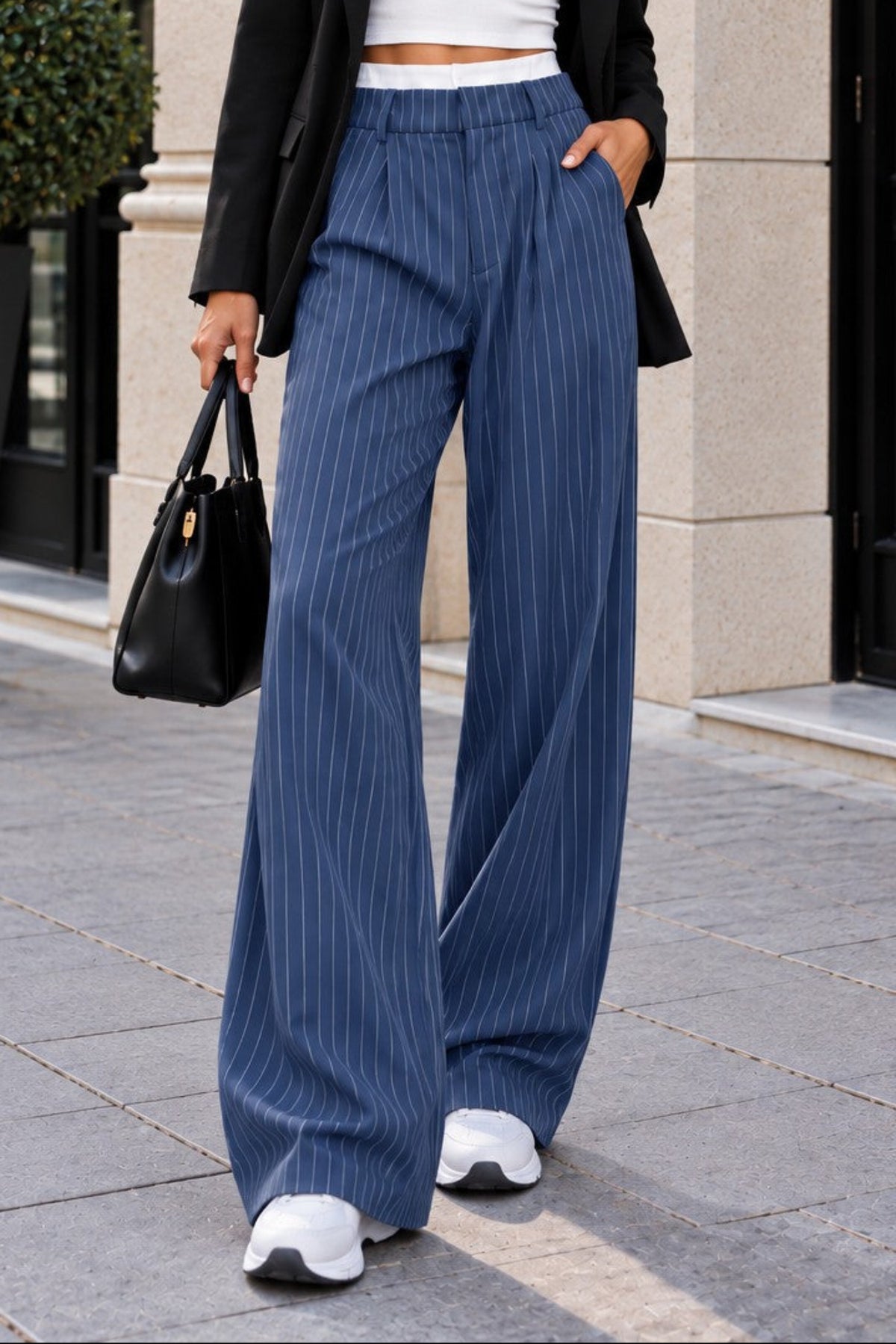 A woman stands on a city sidewalk wearing a black blazer, white crop top, blue pinstriped wide-leg trousers, and white sneakers, while holding a black handbag.