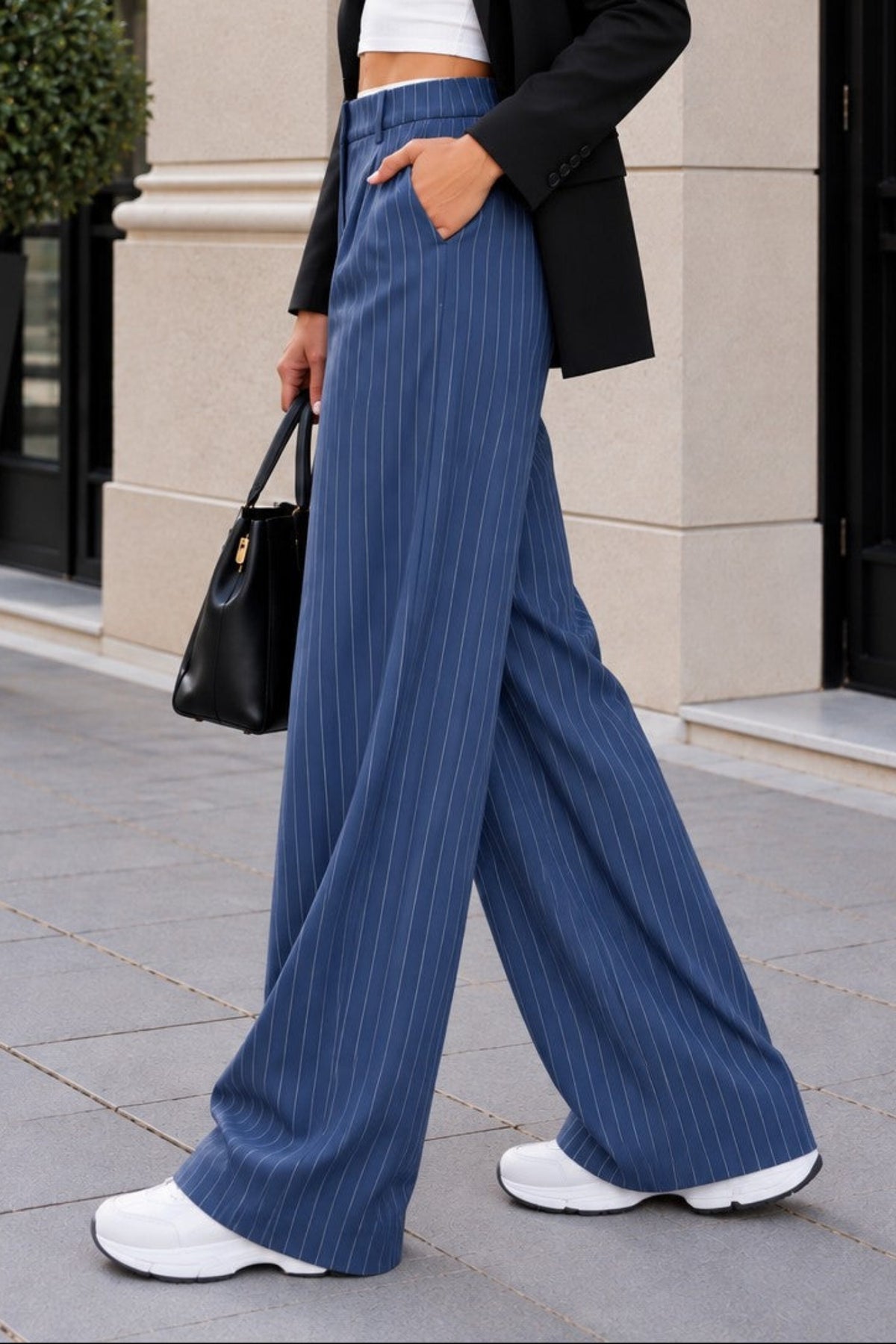 A woman walking outdoors in a city setting wearing a black blazer, white crop top, blue pinstriped wide-leg trousers, white sneakers, and carrying a black handbag.