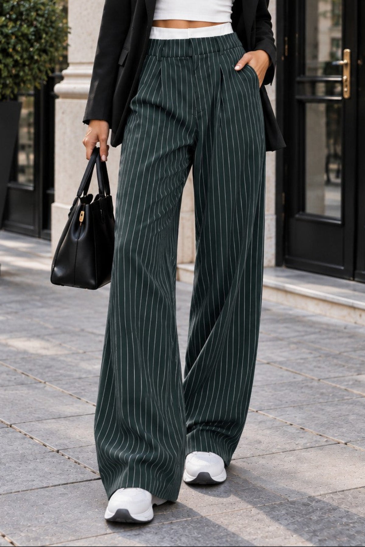 A woman stands on a city sidewalk wearing a black blazer, white crop top, dark green pinstriped wide-leg trousers, and white sneakers, holding a black handbag.