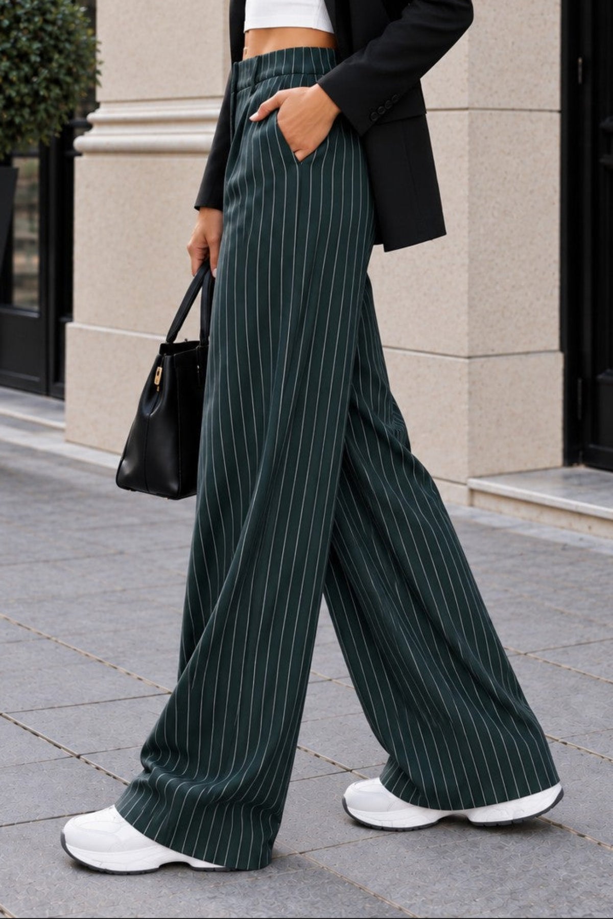 A woman walking outdoors in a city setting wearing dark green pinstripe wide-leg trousers, a white crop top, a black blazer, and white sneakers, holding a black handbag.