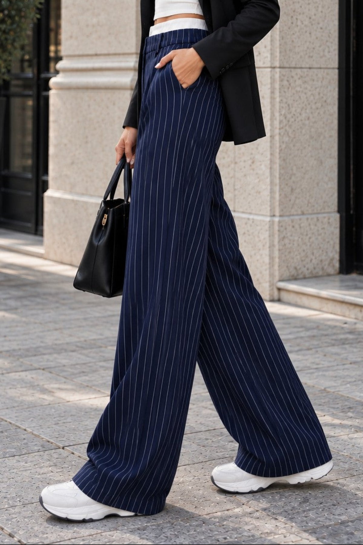 A woman walking outdoors in a city setting wearing a black blazer, white crop top, navy blue pinstripe wide-leg trousers, and white sneakers, holding a black handbag.